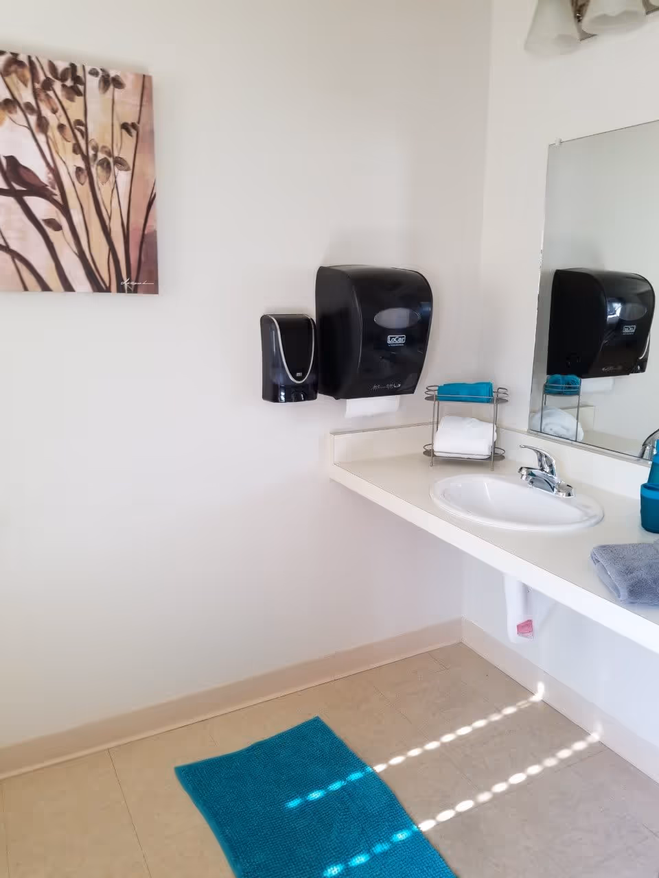 A clean bathroom sink area with a white countertop and a silver faucet. Above the sink is a large mirror reflecting the soap dispenser and paper towel holder mounted on the wall. A small rack holds folded white and teal towels. A teal bath mat is on the floor, and a piece of wall art featuring a bird on branches is hung on the adjacent wall.