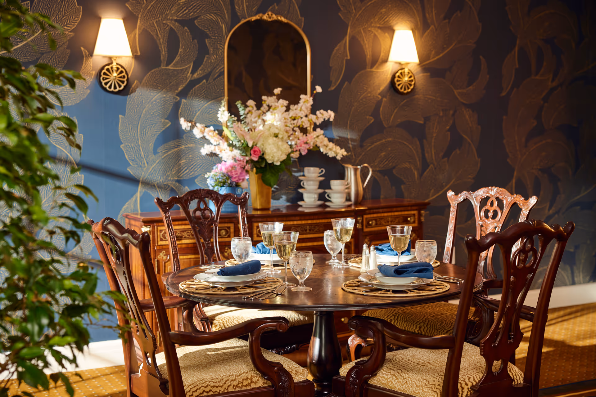 A warmly lit dining room with a round wooden table set for four, featuring elegant place settings with plates, glasses, and blue napkins. The table is surrounded by ornate wooden chairs with cushioned seats. In the background, there is a wooden sideboard with a large floral arrangement, stacked teacups, and a silver pitcher. The walls have a dark blue wallpaper with a large gold leaf pattern, and two wall sconces provide soft lighting.
