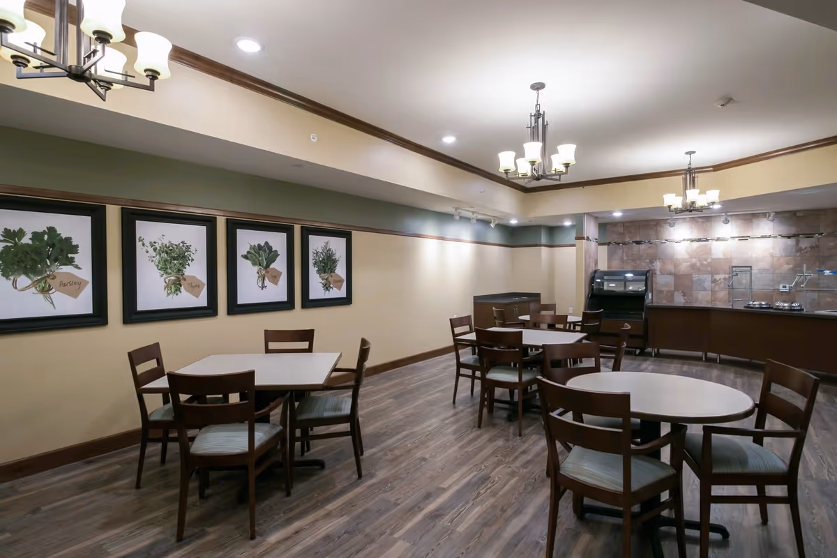 A dining room with several wooden tables and chairs arranged on a wood floor. The walls are painted beige with a green accent stripe and decorated with framed pictures of herbs labeled Parsley, Thyme, Sage, and Rosemary. The ceiling has recessed lighting and hanging light fixtures. In the background, there is a serving counter with a tiled backsplash and a food display case.