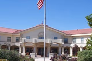 Front entrance of a two-story senior living building with columns, landscaped shrubs, and an American flag on a flagpole.