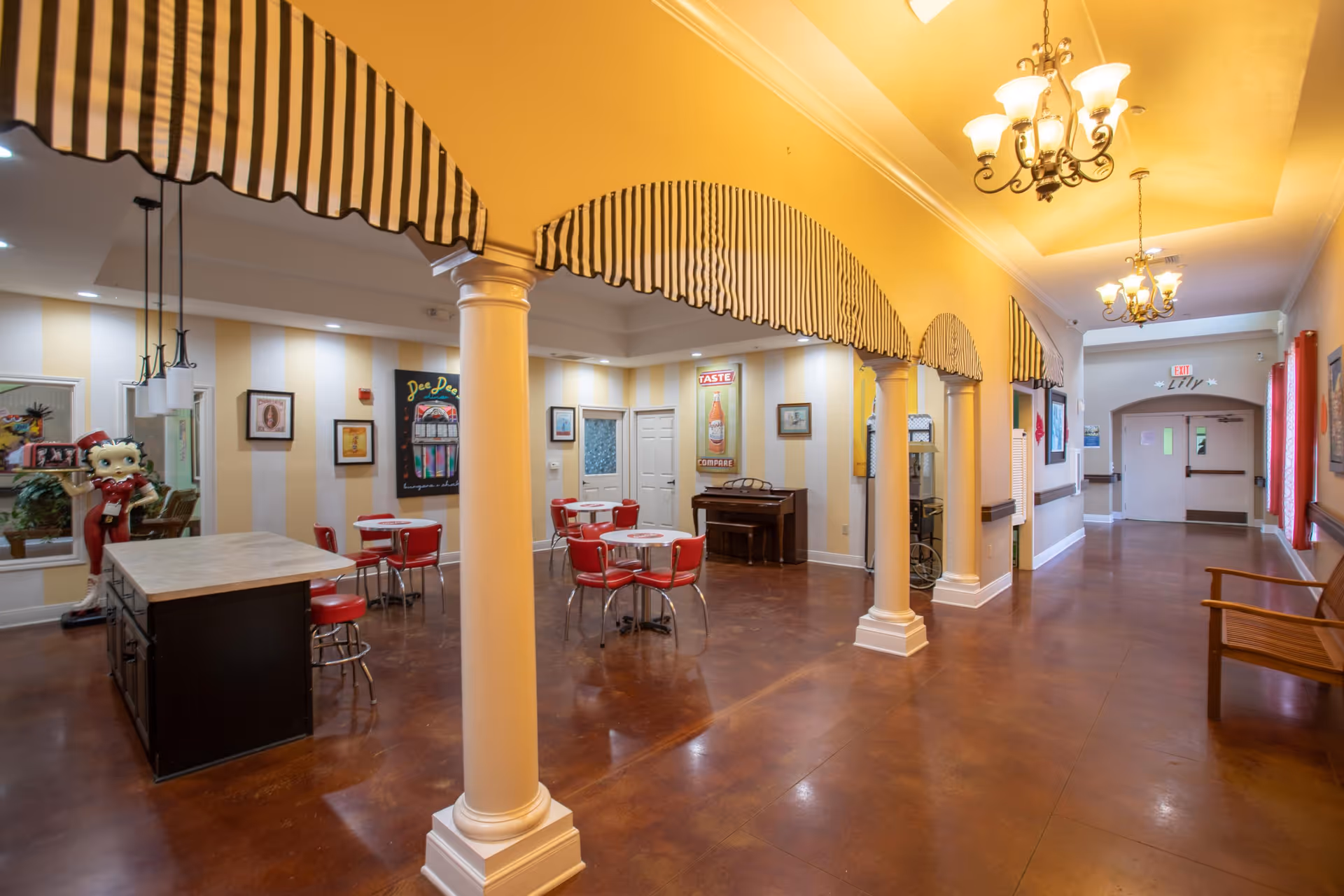 Interior view of a senior living facility common area with yellow walls and striped awnings above white columns. There are several small round tables with red chairs, a piano against the wall, and a statue of Betty Boop near a kitchen island. The floor is polished brown, and chandeliers hang from the ceiling. A hallway with benches and exit signs is visible in the background.