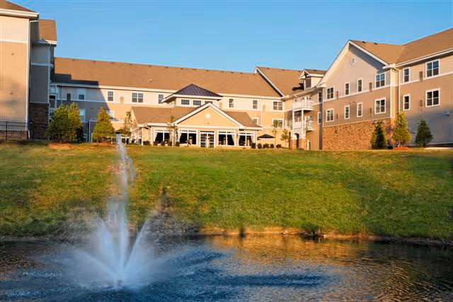 Exterior view of Pinebrook of Milford by StoryPoint Group, showing a multi-story senior living facility building with beige siding and brick accents. In the foreground, there is a pond with a water fountain spraying water upwards, surrounded by a grassy area and some small trees under a clear blue sky.