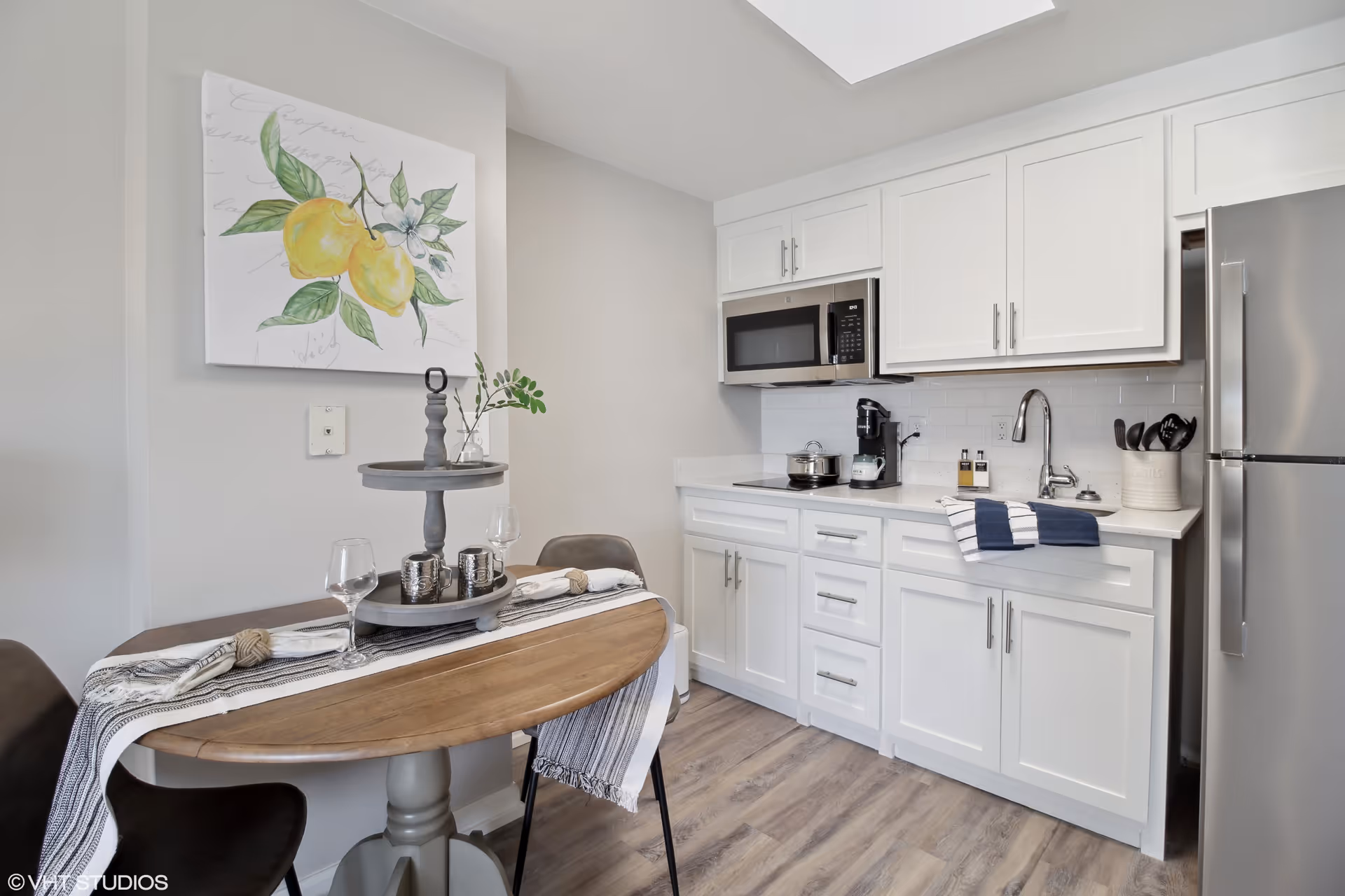 A small modern kitchen with white cabinets, a stainless steel refrigerator, microwave, and sink. A wooden dining table with a gray two-tiered tray, two wine glasses, and a striped table runner is set against the wall. Above the table is a painting of lemons with green leaves on a white background. The floor is light wood.