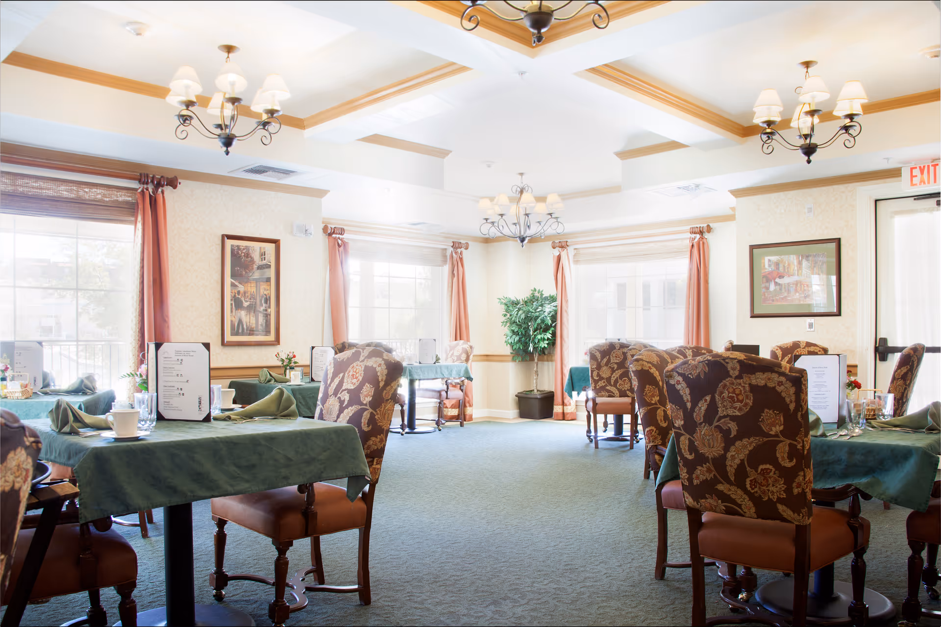 Sunlit dining room with multiple tables set with green tablecloths, napkins, and upholstered chairs.
