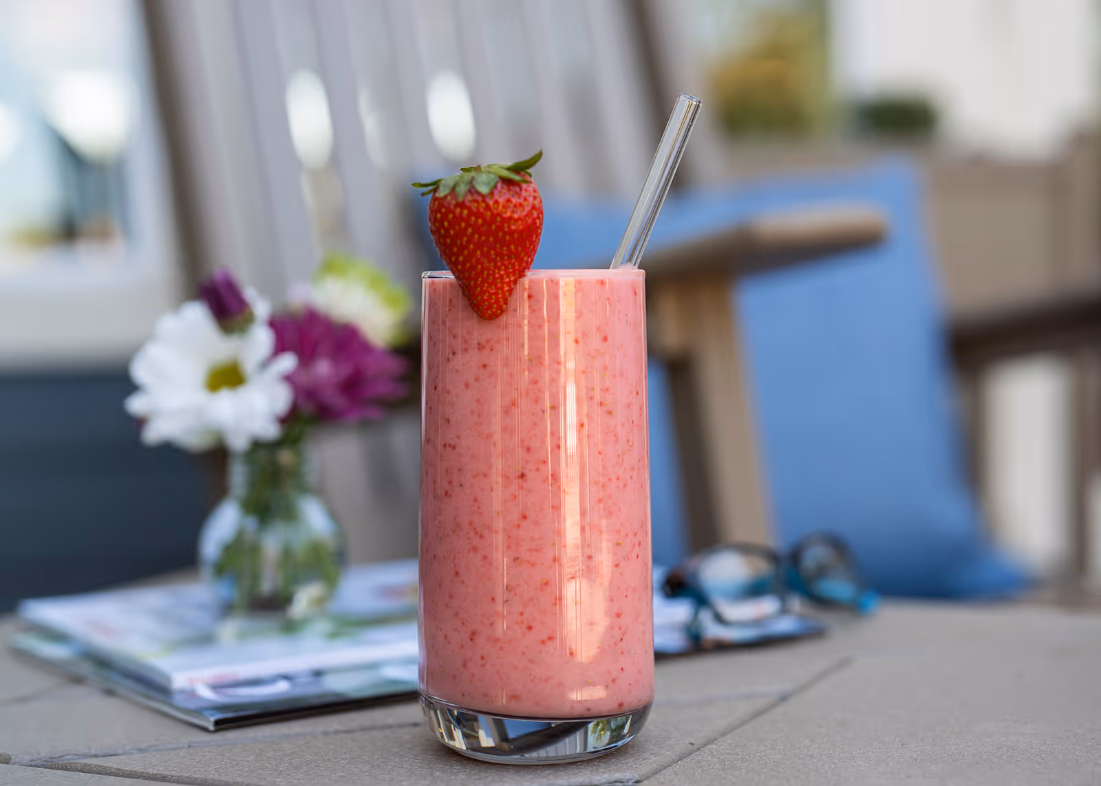 A tall glass of pink strawberry smoothie with a whole strawberry on the rim and a clear straw, placed on a table. In the background, there is a small vase with white and purple flowers, a pair of glasses, and some magazines on the table, with blurred outdoor furniture and cushions.