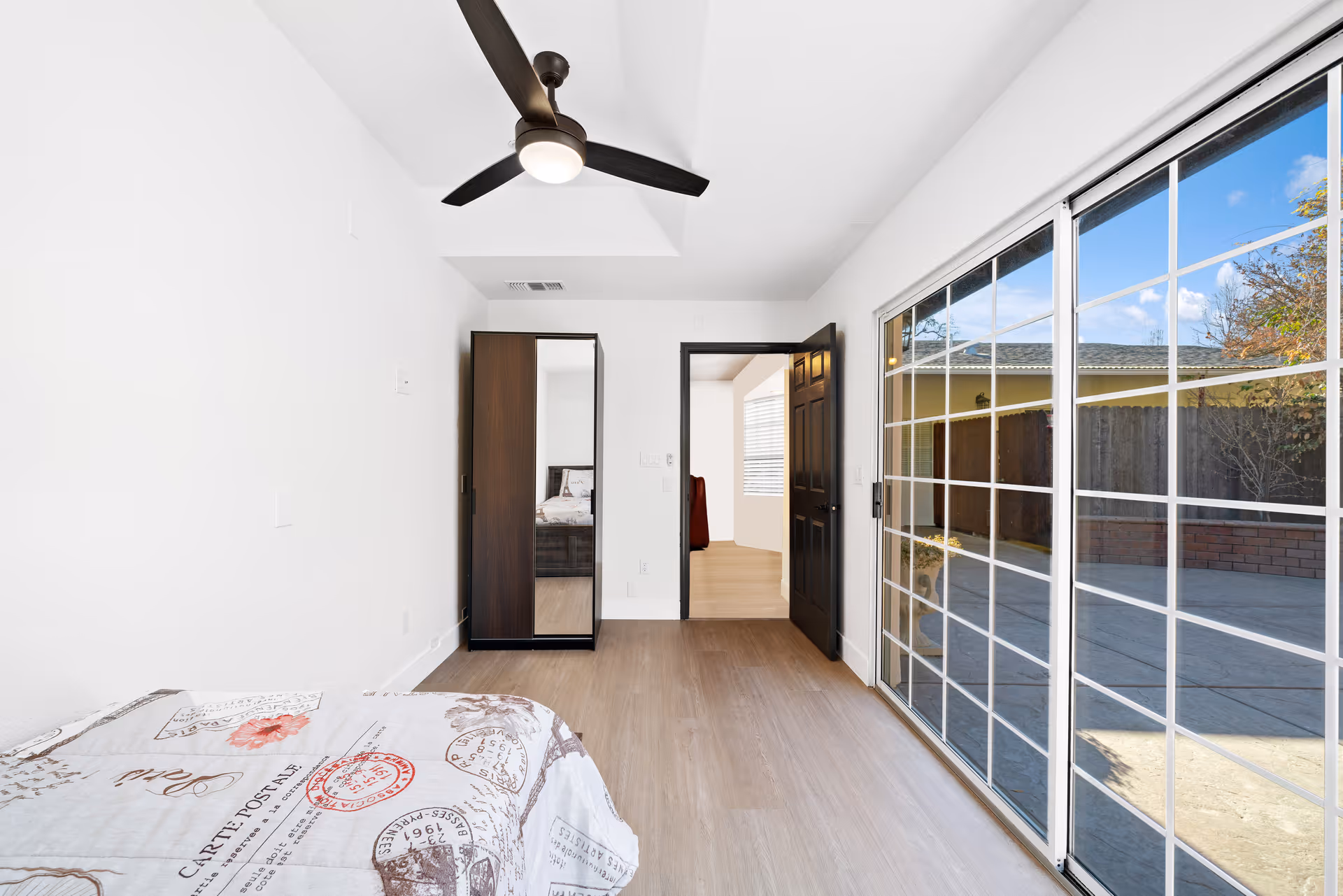 A bright bedroom with a bed partially visible on the left, a ceiling fan with light, a wardrobe with a mirrored door, and a sliding glass door leading to an outdoor patio area. The room has light-colored walls and wood flooring.