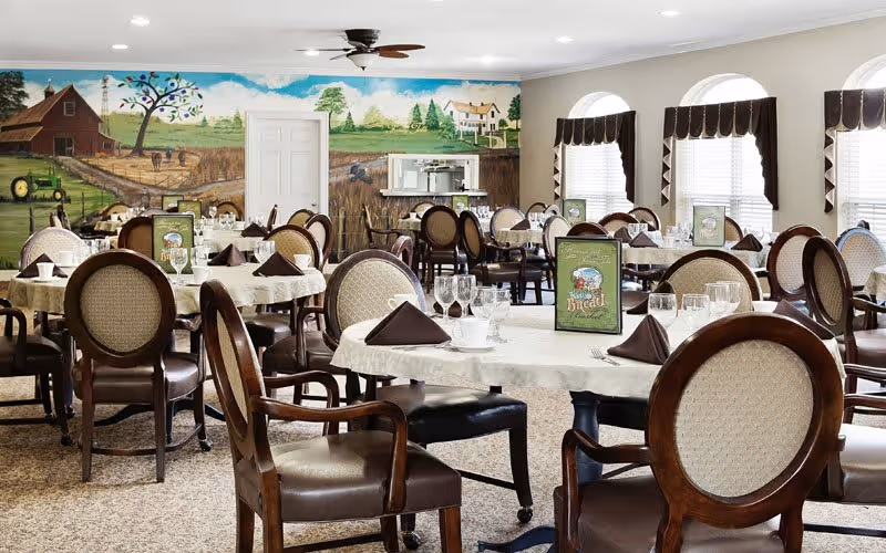 A bright and spacious dining room with multiple round tables covered in white tablecloths, each set with glassware, cups, and brown folded napkins. The chairs have wooden frames with cushioned seats and backs. The room features large windows with brown valances, and a mural on one wall depicting a farm scene with a barn, tractor, trees, and fields under a blue sky. A ceiling fan is mounted above.