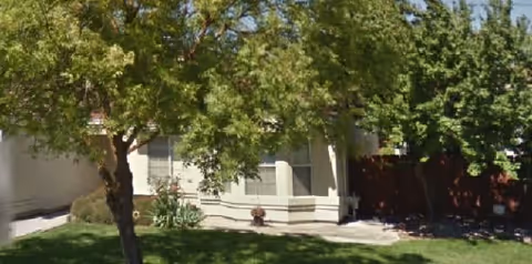 Exterior view of a single-story residential building partially obscured by trees with green leaves. There is a well-maintained lawn in front and a concrete pathway leading to the entrance.