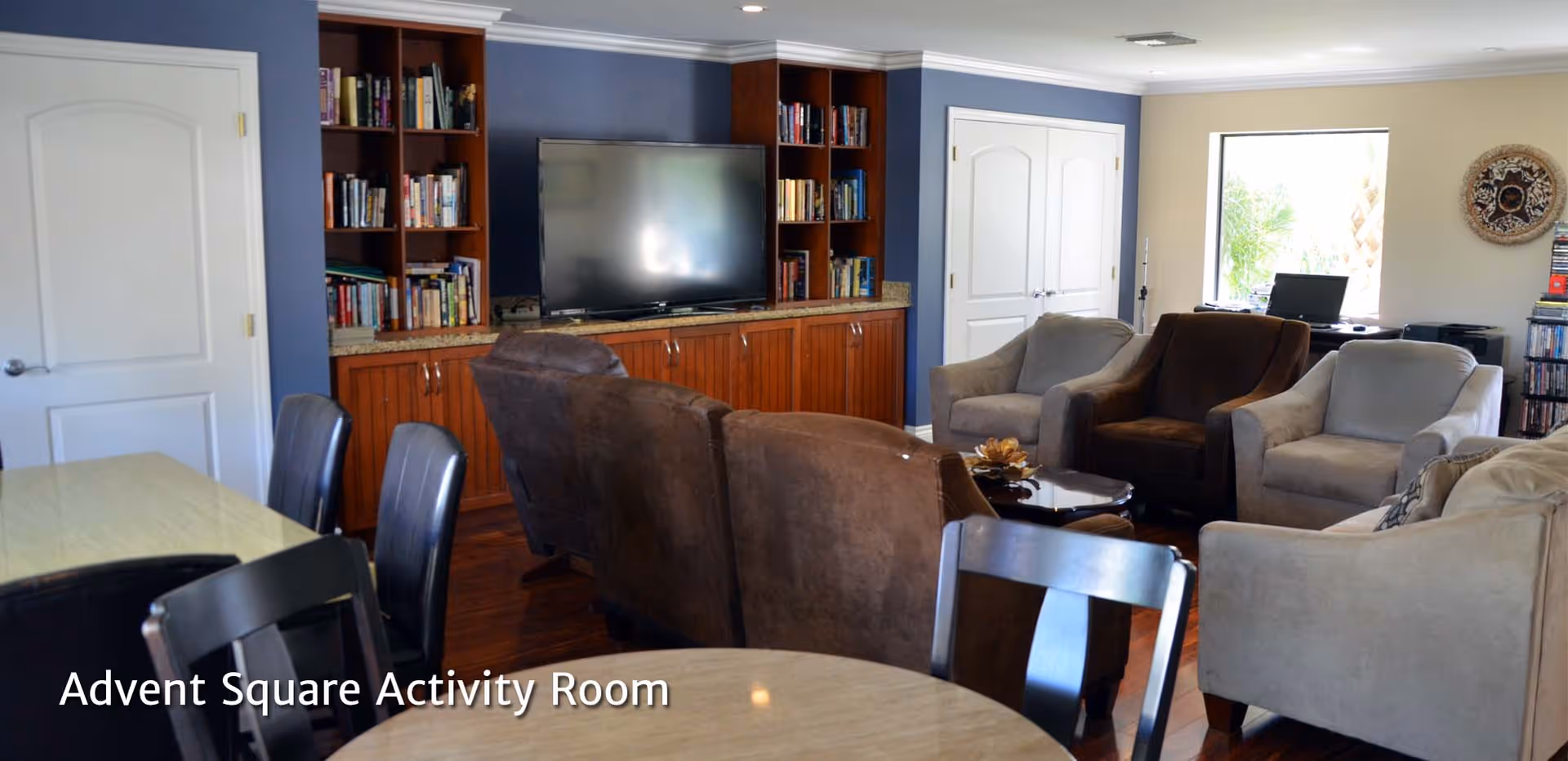 A cozy activity room with multiple armchairs and a sofa arranged around a small coffee table. There is a large flat-screen TV mounted on a wooden cabinet with built-in bookshelves filled with books. The room has wooden flooring, a window letting in natural light, and a table with chairs in the foreground.