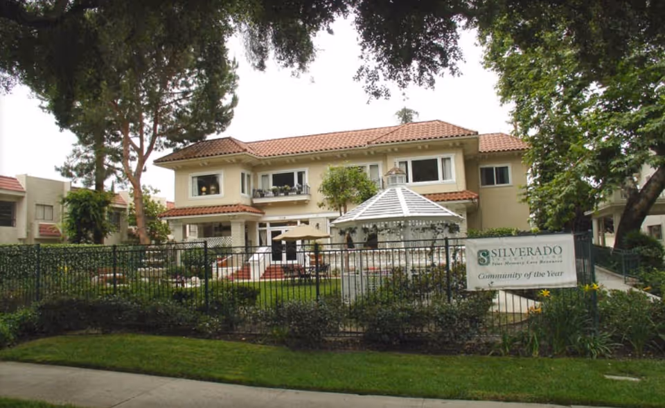 Exterior view of Silverado The Huntington Memory Care Community building with a fenced garden area, a white gazebo, trees, and a sign that reads 'Silverado The Memory Care Resource Community of the Year'.
