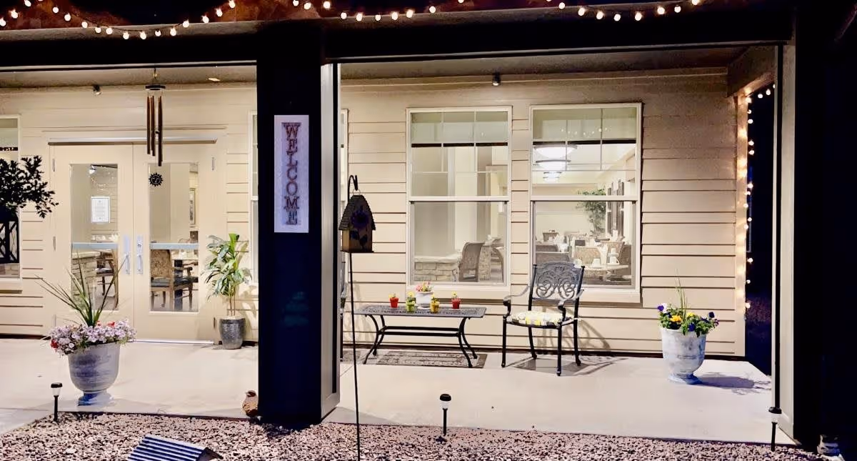Outdoor patio area at night with a welcome sign on a pillar, two potted plants with flowers, a metal bench with small flower pots on it, and a metal chair with a cushion. Behind the patio are large windows and double glass doors showing a well-lit interior room with tables and chairs.