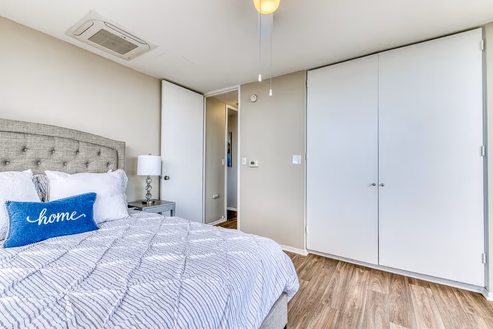 A bright bedroom with a gray upholstered headboard bed, white and blue bedding including a blue pillow with the word 'home' on it, a bedside table with a lamp, white walls, a ceiling fan, a door slightly open leading to a hallway, and a large white closet with double doors.