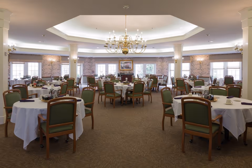 A spacious dining room with round tables covered in white tablecloths, each surrounded by green cushioned wooden chairs. The room features a large chandelier hanging from a recessed ceiling, floral wallpaper, and multiple windows allowing natural light to fill the space.