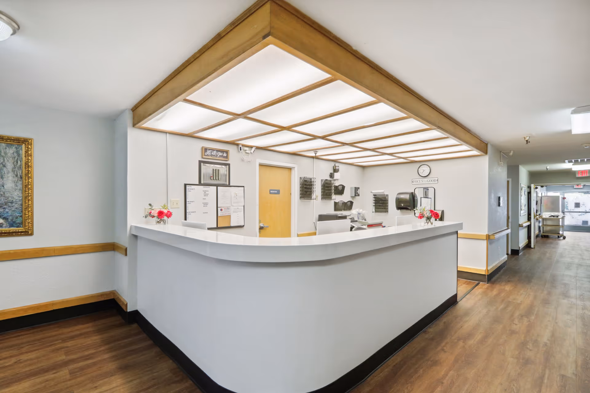 Reception desk area inside a healthcare facility with a white curved counter, overhead wooden framed lighting, a bulletin board, a door, and a hallway leading further into the building. The floor is wood, and there are small flower arrangements on the counter.
