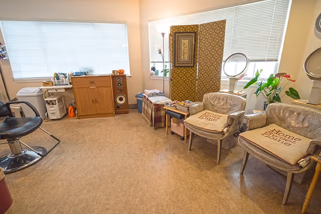 A cozy salon or beauty room with two chairs covered in plastic and cushions that say 'Home Sweet Home', a black salon chair, a wooden cabinet with speakers, a small table with various items, a decorative folding screen, and a large window with blinds letting in natural light.