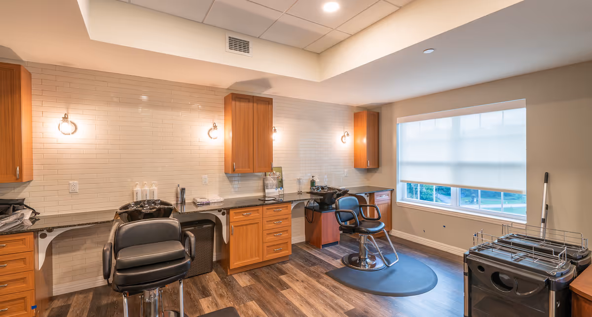 Interior of a hair salon area in a senior living facility with two black salon chairs, sinks for washing hair, wooden cabinets, a large window with a white roller shade, and wood flooring.