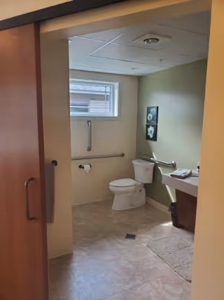 A clean, accessible bathroom with a toilet, grab bars on the walls, a small window, a sink with a countertop, and a trash bin. The floor is tiled and there is a beige rug near the sink. Two framed floral pictures hang on the wall above the toilet.