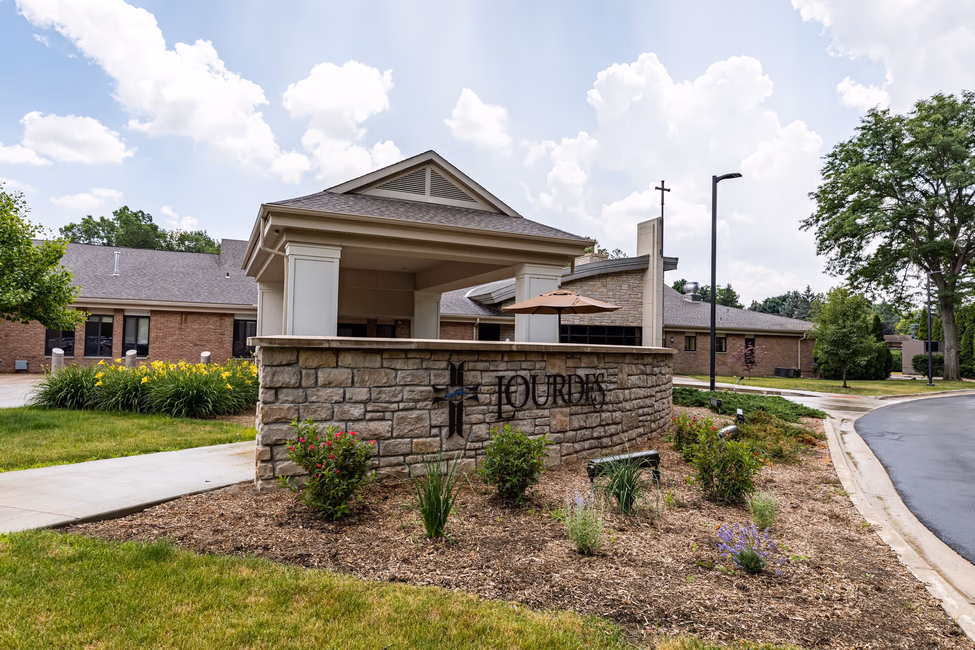 Front entrance of Lourdes Senior Community showing a stone sign, covered portico, and landscaped beds.