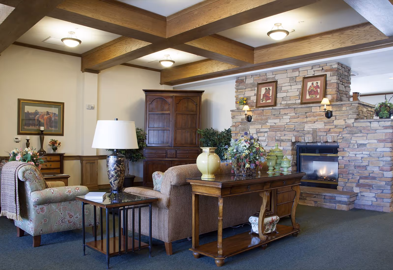 A cozy living room area with wooden ceiling beams, a stone fireplace with two framed pictures and wall sconces above it, a wooden cabinet, a floral armchair with a throw blanket, a beige sofa, a wooden console table with decorative vases and a flower arrangement, and a side table with a lamp and a book.