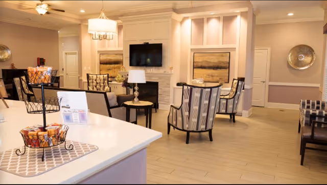 Bright, furnished assisted-living common room with chairs and small tables facing a wall-mounted TV above a fireplace, and a countertop with decorative baskets in the foreground.
