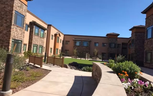 Sunlit landscaped courtyard with a paved walkway, flower beds, and a surrounding two-story senior living building under a clear blue sky.