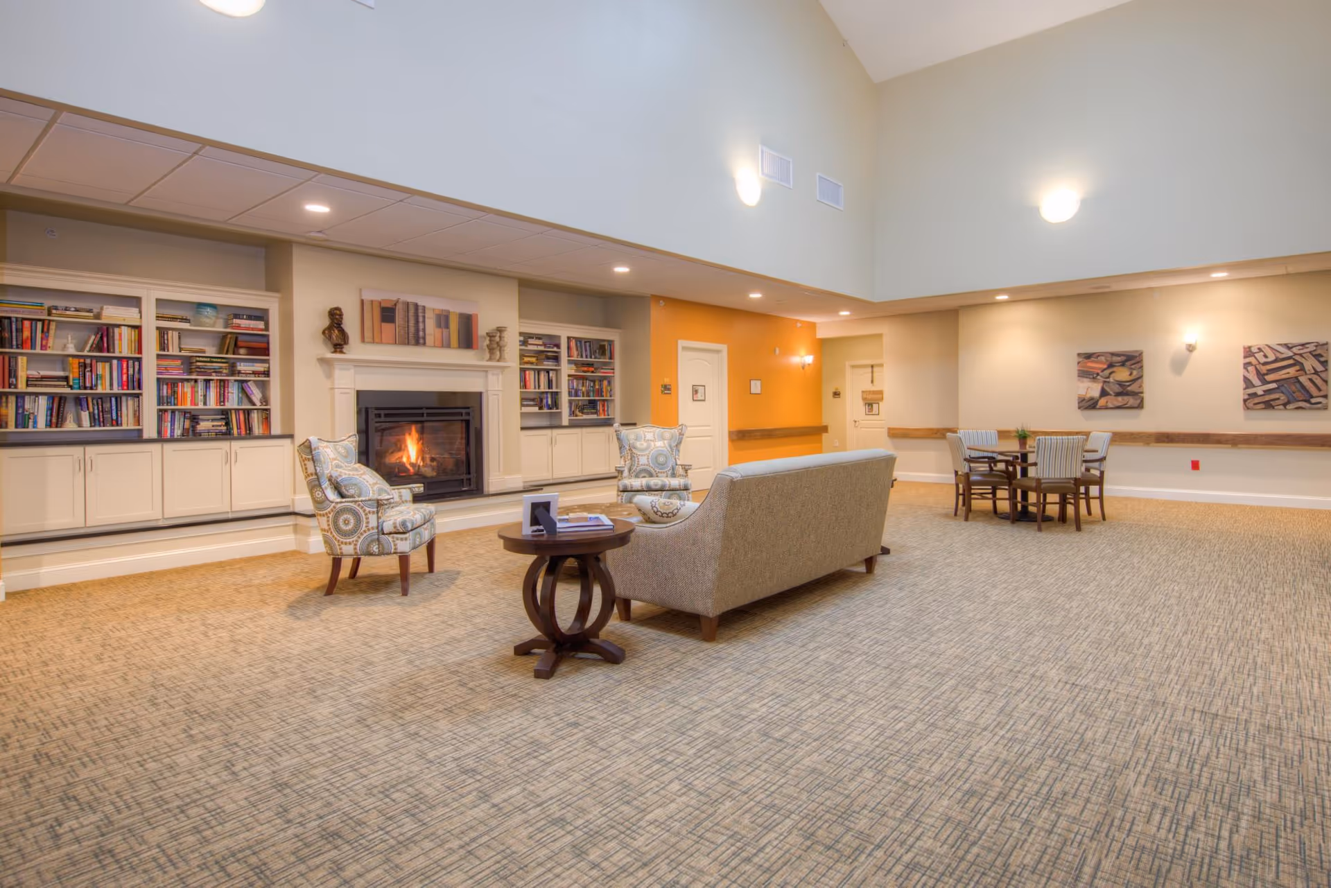 A spacious living room area with a high ceiling, featuring a fireplace surrounded by built-in bookshelves filled with books. In front of the fireplace, there are two patterned armchairs and a beige sofa with a round wooden side table. The room has carpeted flooring, warm lighting, and a small dining table with four chairs in the background. The walls are painted in neutral tones with one accent wall in orange.