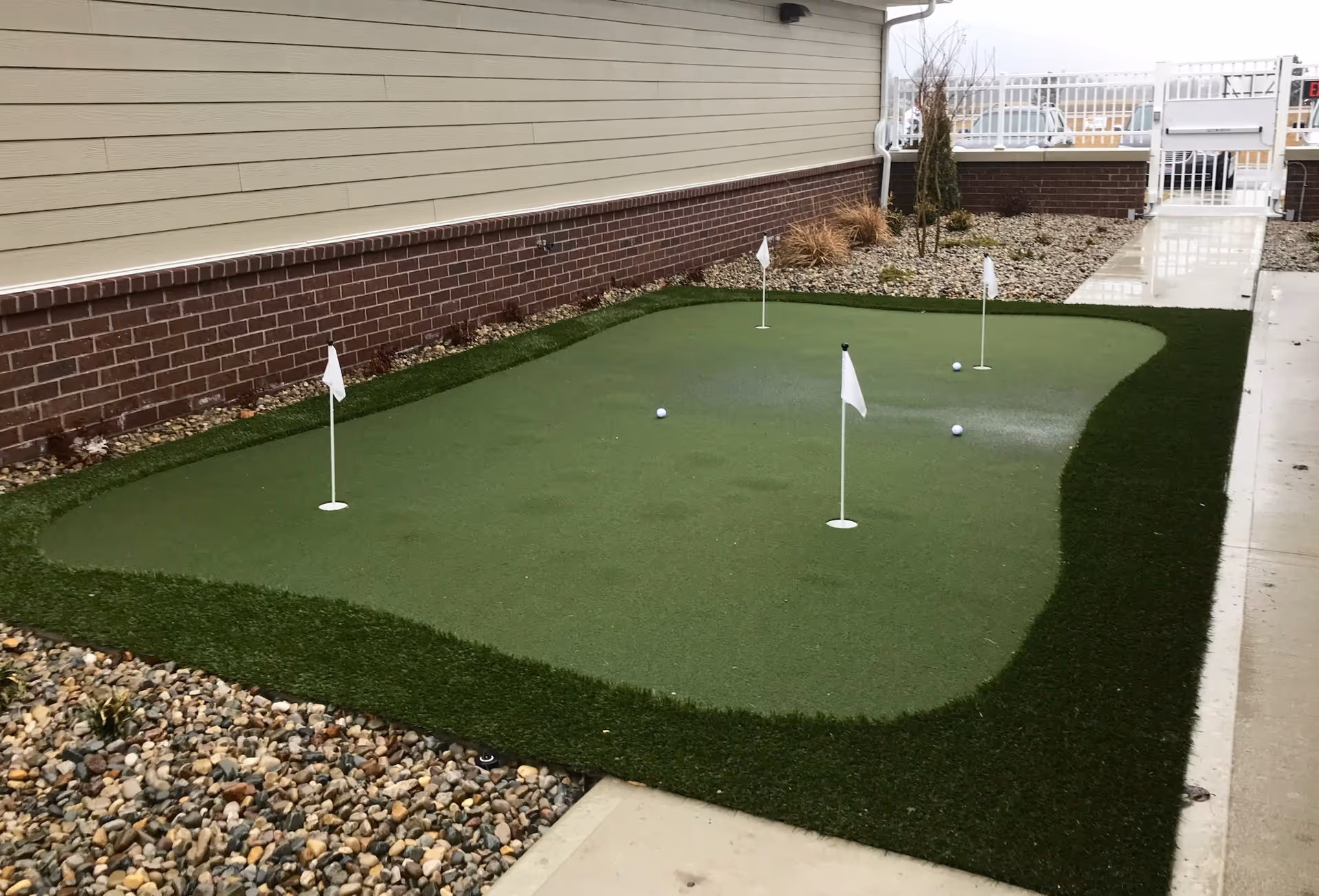 Outdoor putting green with four small white flags and several golf balls, surrounded by a border of artificial turf and landscaping rocks, next to a building with beige siding and red brick lower wall.