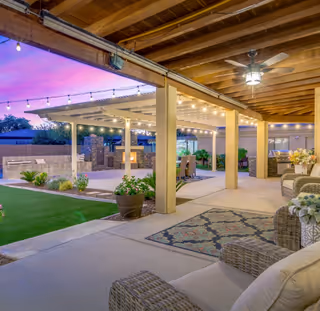 Covered outdoor patio with wicker seating, string lights, ceiling fan, dining area and fireplace overlooking a landscaped courtyard at sunset.