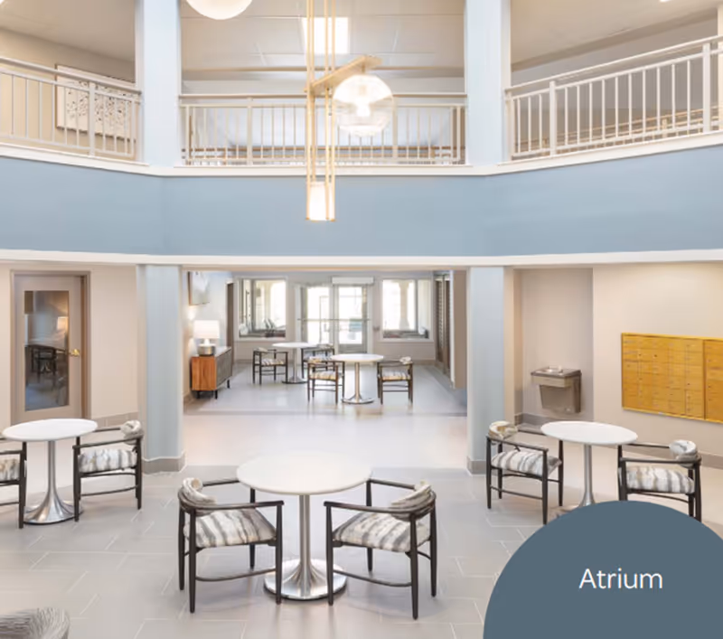 Bright and spacious atrium area with round tables and cushioned chairs arranged for seating. The space features a high ceiling with a hanging light fixture, a second-floor balcony railing, and a wall of mailboxes on the right side. Large windows and doors at the back allow natural light to fill the room.