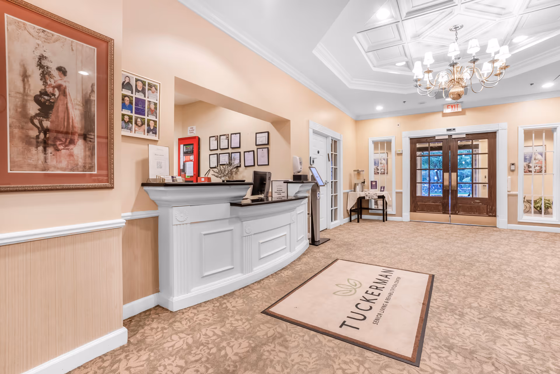 Reception area of The Terraces at Tuckerman Lane featuring a white front desk with a computer, a large framed painting on the wall, several framed photos and certificates, a chandelier hanging from a coffered ceiling, double wooden doors with glass panels, and a floor mat with the Tuckerman logo.