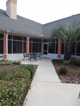 Outdoor courtyard area with a concrete walkway leading to a small patio set with a table and four chairs. The patio is surrounded by low bushes and a palm tree, with a building featuring screened windows and a sloped roof in the background.