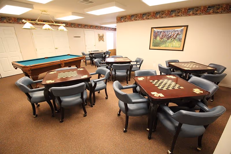 A recreational room with several square tables featuring built-in checkerboards and card holders, surrounded by blue cushioned chairs. In the background, there is a pool table and more seating. The walls are beige with a framed picture of horses racing and a decorative border near the ceiling.