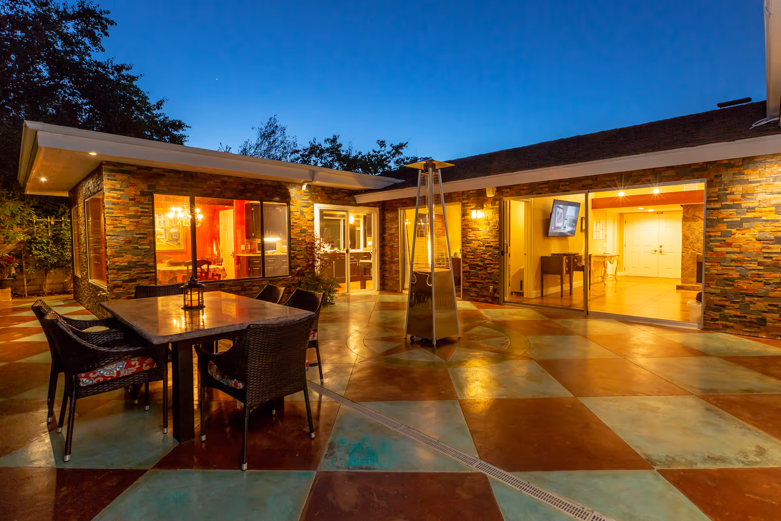 Outdoor patio area at dusk with a square table surrounded by six wicker chairs with patterned cushions. The patio floor has a colorful geometric tile pattern. The building exterior features stone walls and large windows with warm interior lighting visible inside. A tall outdoor heater stands near the table.