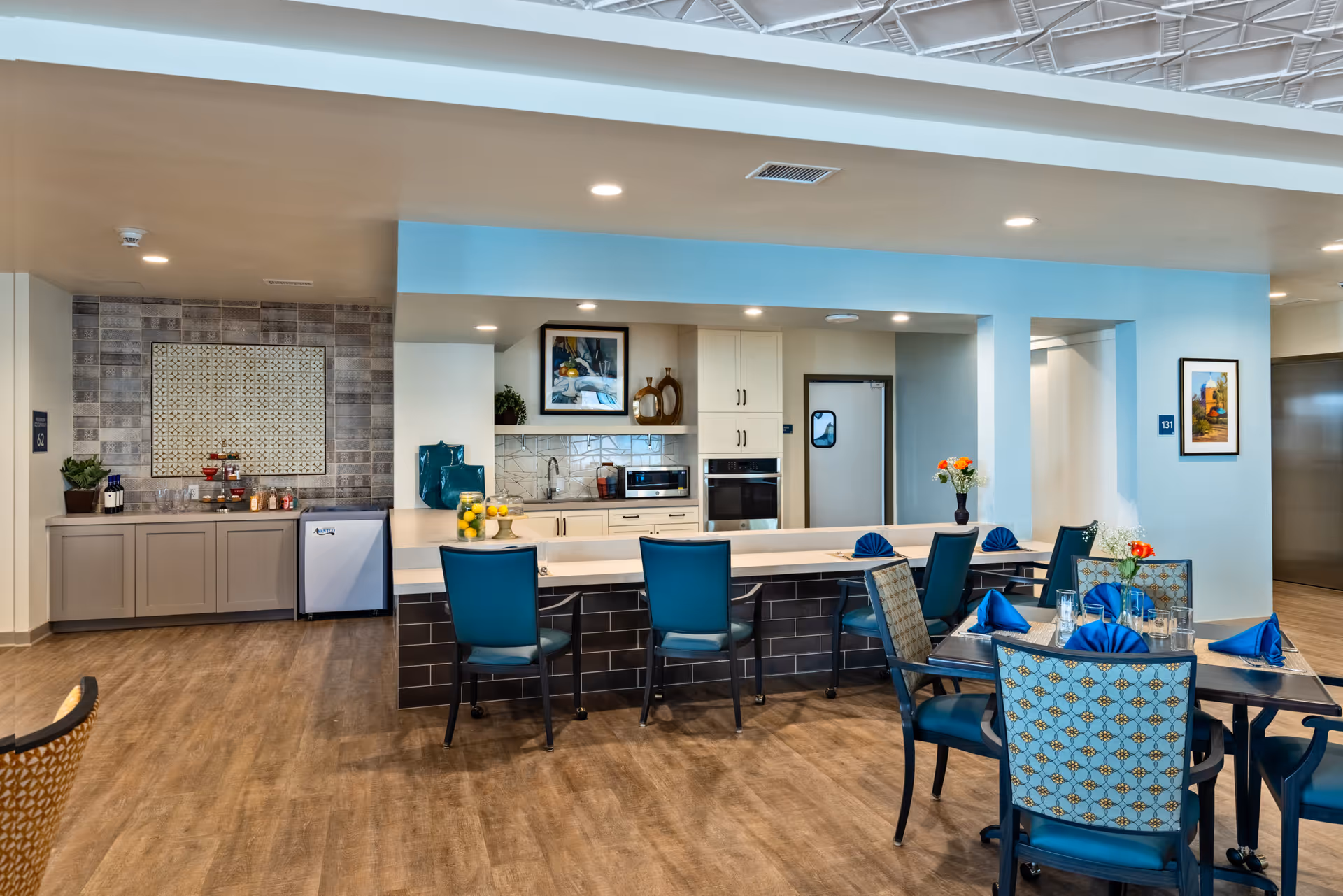 A modern dining and kitchen area in a senior living facility with a long counter and four teal chairs. The kitchen features white cabinets, a microwave, and a refrigerator. There are tables with blue napkins and glasses set for dining. The floor is wood-style, and the walls are light-colored with some artwork and decorative items.