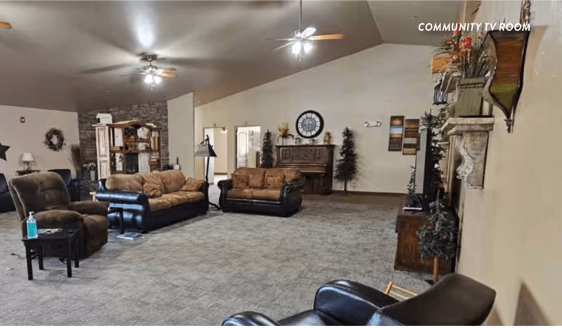A spacious community TV room with multiple brown and black leather sofas and armchairs arranged around the room. The room features a stone fireplace, a wooden cabinet with a large clock above it, decorative plants, and ceiling fans with lights. The walls are painted beige, and the carpet is gray. The words 'COMMUNITY TV ROOM' are visible on the upper right corner of the image.