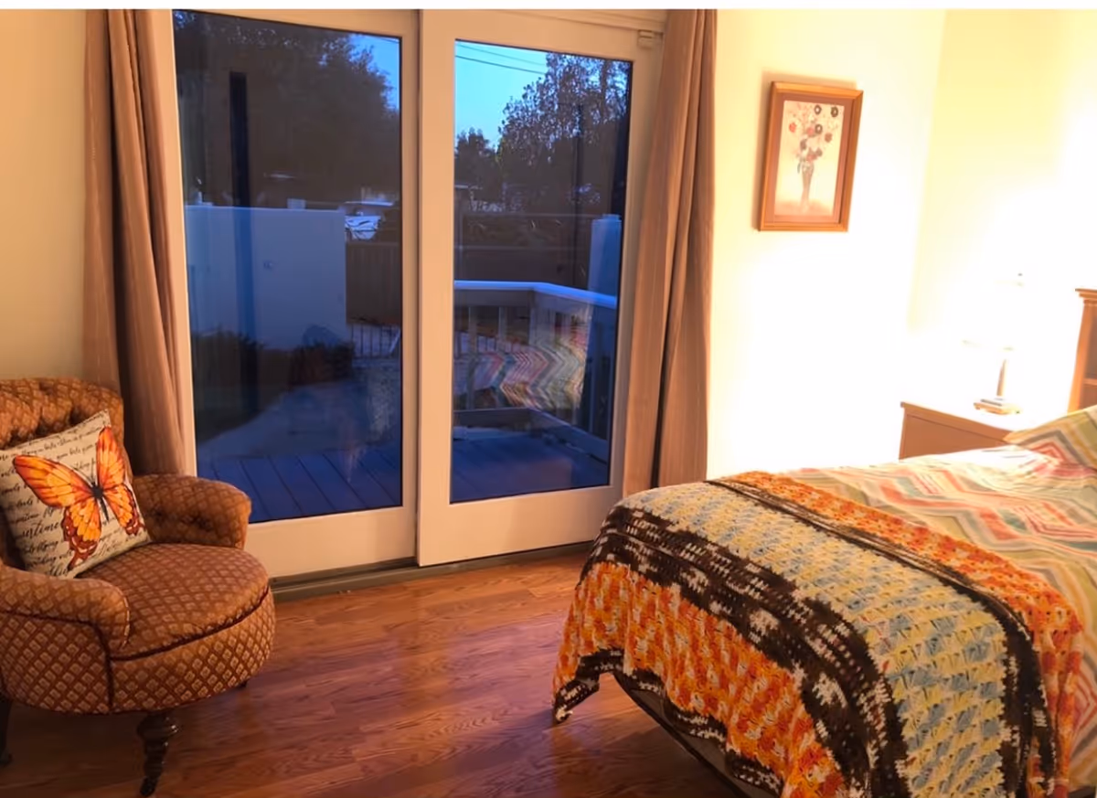 Cozy bedroom with wooden floor, a patterned armchair with a butterfly pillow, a bed with a colorful crocheted blanket and chevron-patterned bedding, a nightstand with a lamp, a framed floral picture on the wall, and large glass sliding doors leading to an outdoor deck.