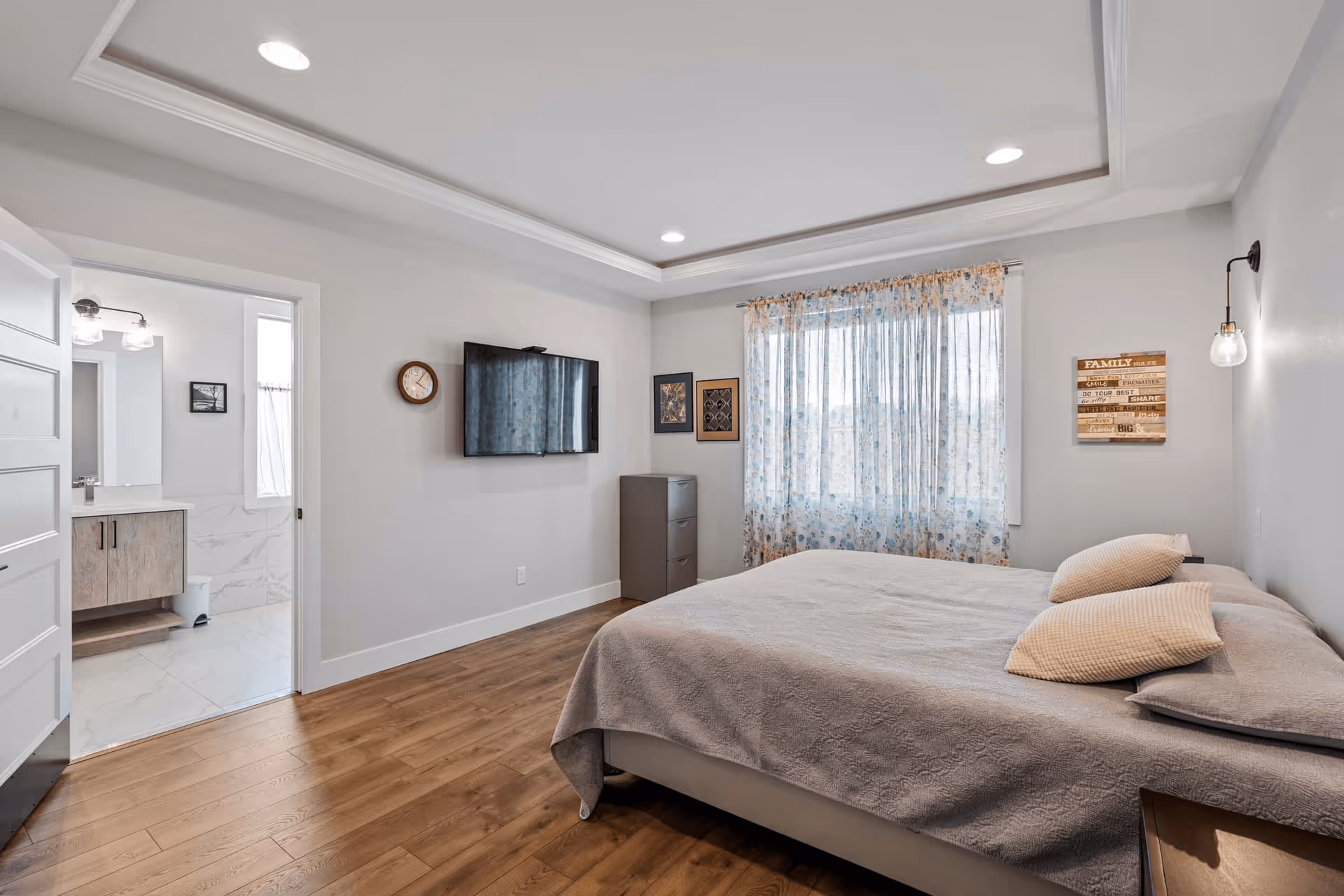 A modern bedroom with a large bed covered in a light gray bedspread and two pillows. The room has wooden flooring, a wall-mounted flat-screen TV, a small chest of drawers, and a window with sheer floral curtains. A door on the left leads to a bathroom with a vanity and mirror visible. The walls are painted light gray, and there is a decorative wall light and framed artwork on the walls.