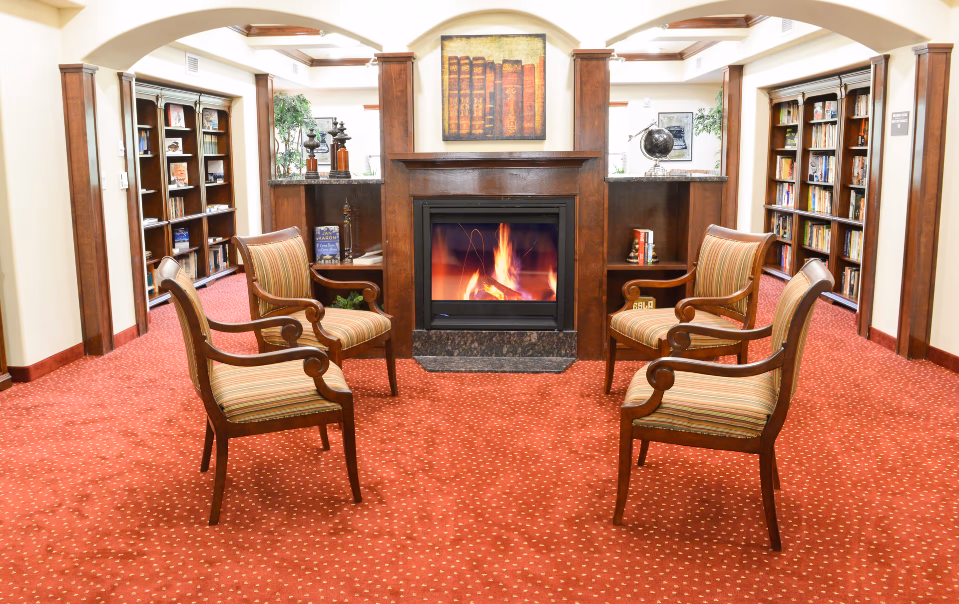 A cozy sitting area with four wooden armchairs arranged around a fireplace with a burning fire. The room has red carpet with a dotted pattern, wooden bookshelves filled with books on either side, and decorative items including a globe and framed artwork above the fireplace.