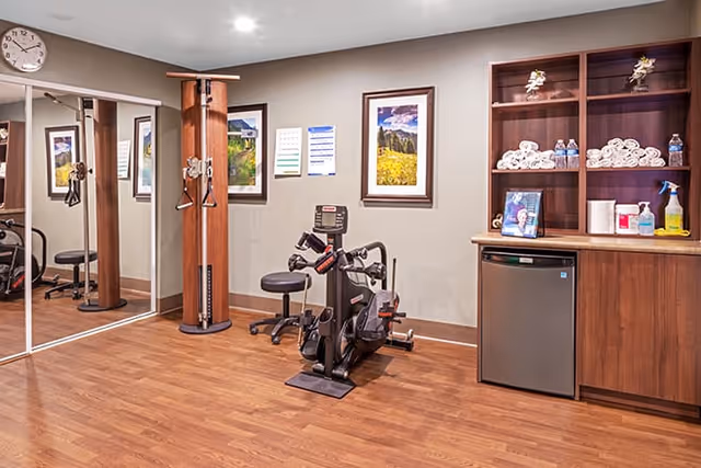 A small fitness room with wooden flooring featuring exercise equipment including a stationary bike and a multi-use pulley machine. There is a large mirror on one wall, framed landscape pictures on another wall, and a wooden cabinet with shelves holding rolled towels, water bottles, cleaning supplies, and a mini refrigerator.