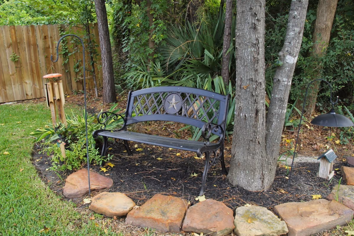 A black metal garden bench with a star design on the backrest is placed on a mulched area surrounded by large stones. The bench is next to a tree and surrounded by green plants and a wooden fence in the background. There are two hanging garden decorations on metal hooks nearby.