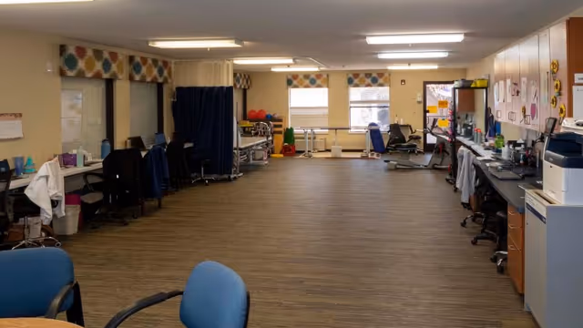 A spacious rehabilitation room with exercise equipment, chairs, desks, and medical supplies. The room has large windows with patterned valances, wood flooring, and fluorescent ceiling lights.