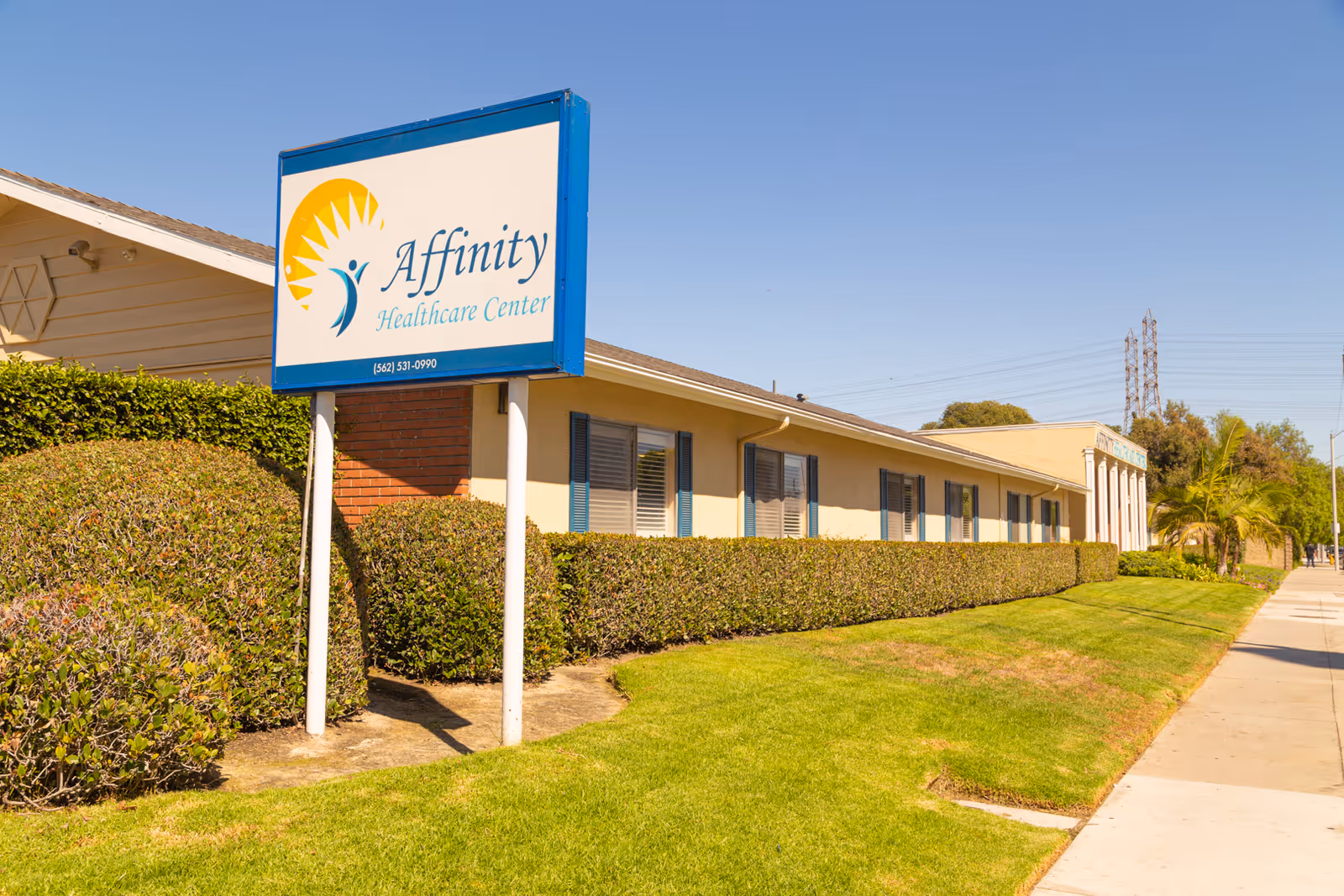 Exterior view of a single-story healthcare center building with beige walls and blue window shutters. A large sign in front reads 'Affinity Healthcare Center' with a sun and person logo. The building is surrounded by trimmed bushes and a green lawn under a clear blue sky.
