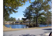 A peaceful outdoor scene featuring a paved walking path alongside a calm body of water with trees and greenery surrounding it. A white bench is placed near the water's edge, and buildings are visible in the background across the water under a clear sky.