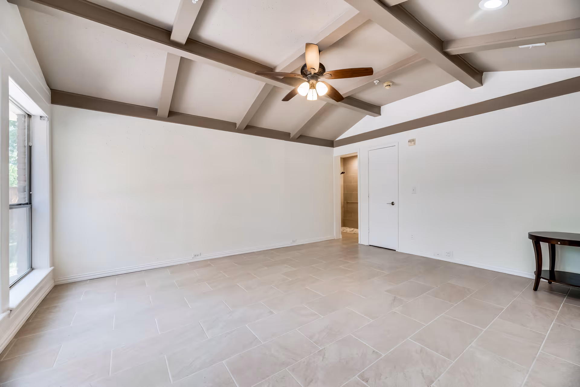 Spacious empty living room with a vaulted beamed ceiling, ceiling fan, large window, and tiled floor.