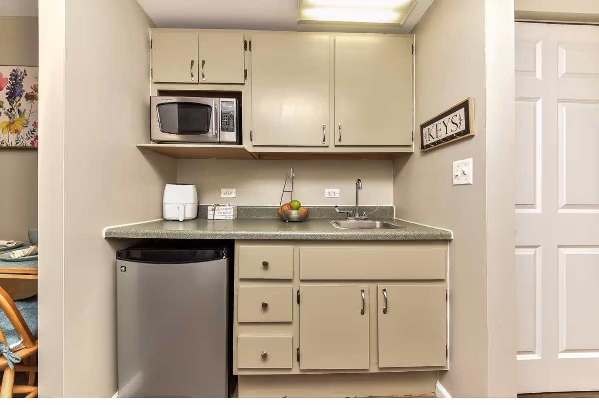 Small kitchen area with beige cabinets, a microwave, a mini refrigerator, a sink, and a countertop with a bowl of fruit and a small appliance. A sign labeled 'KEYS' is mounted on the wall next to a white door.