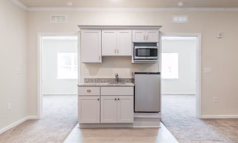 Small kitchenette area with white cabinets, a granite countertop, a sink, a microwave, and a mini refrigerator. The kitchenette is positioned between two doorways leading to carpeted rooms with windows letting in natural light.