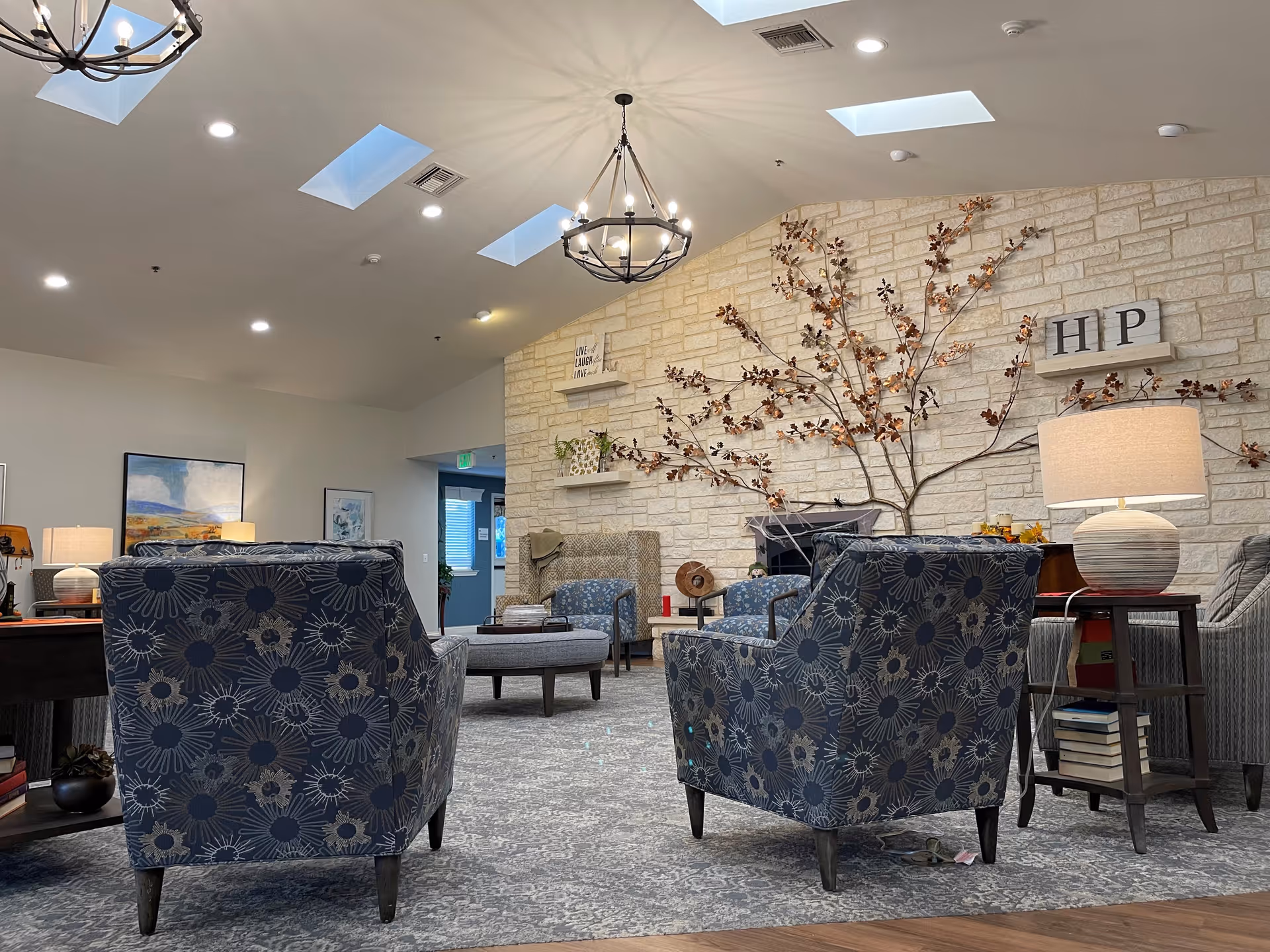 A cozy living room area with patterned armchairs, a stone wall decorated with a metal tree sculpture, and shelves holding decorative items. The room has a high ceiling with skylights and a chandelier, a carpeted floor, and a side table with a lamp and books.