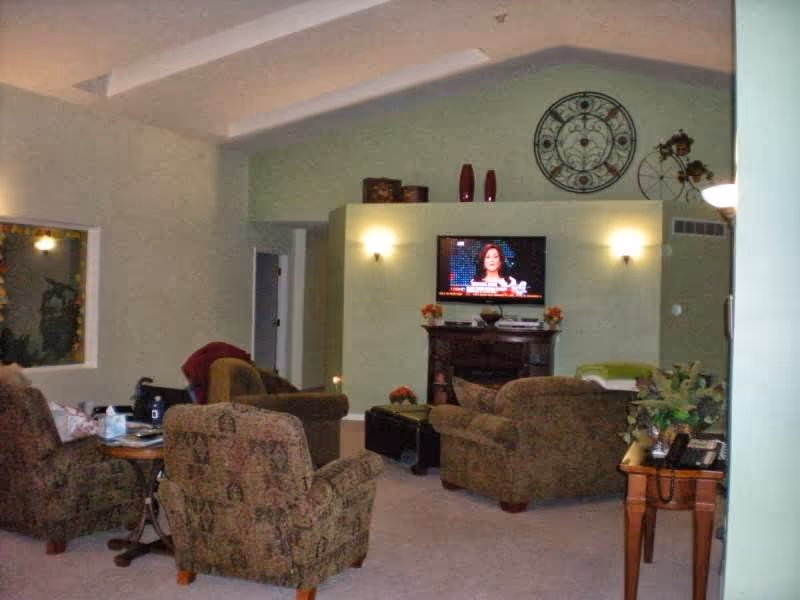 Communal living room with upholstered chairs and sofas arranged around a TV mounted on a decorative wall beneath high ceilings.