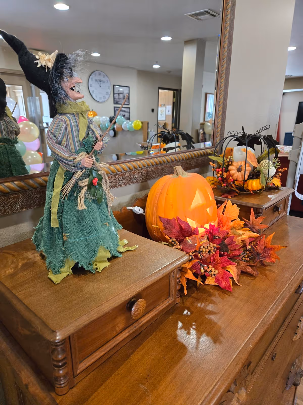 A wooden dresser topped with Halloween decorations including a witch doll, a lit jack-o'-lantern, and autumn foliage in front of a large mirror reflecting the facility interior.