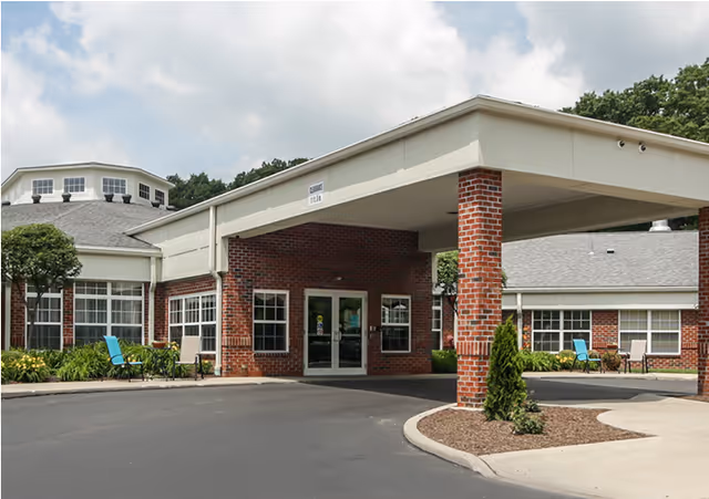 Entrance of a senior living facility with a covered drop-off area supported by brick columns, surrounded by a paved driveway and landscaped with small bushes and outdoor seating.